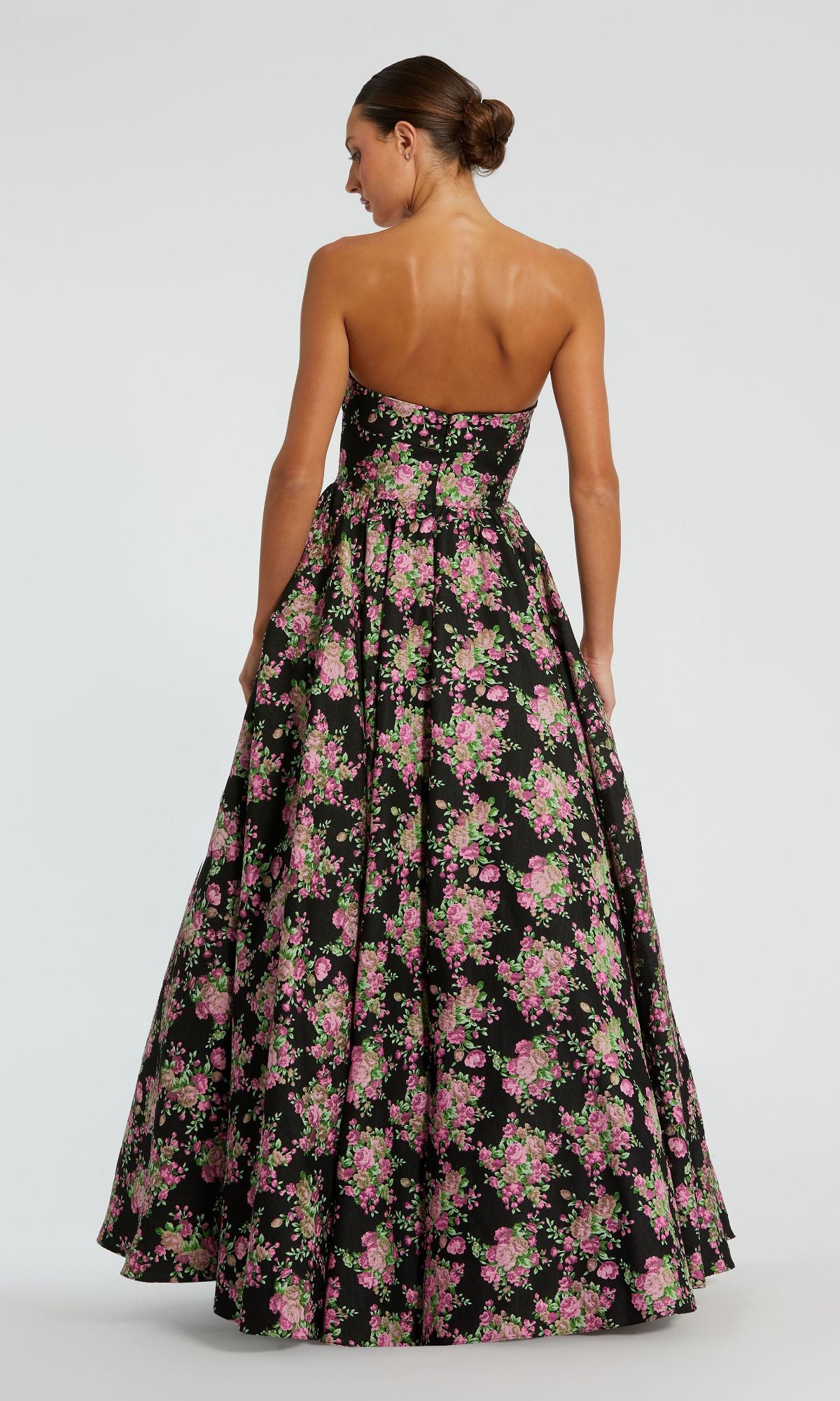 A woman faces away, wearing the Mac Duggal 68780 Strapless Print Formal Dress featuring a pink and green floral design. The floor-length A-line gown suits special occasions, and her hair is styled neatly in a bun.